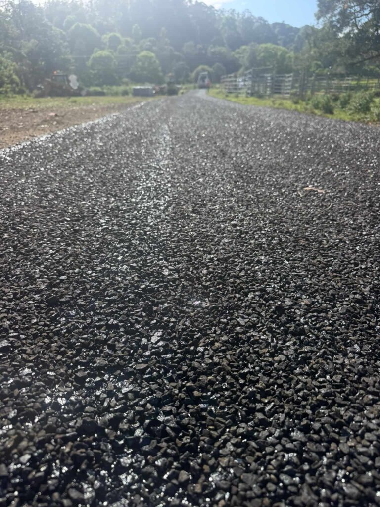 A freshly spray-sealed asphalt road with loose dark aggregate in the foreground, a road roller working in the distance.